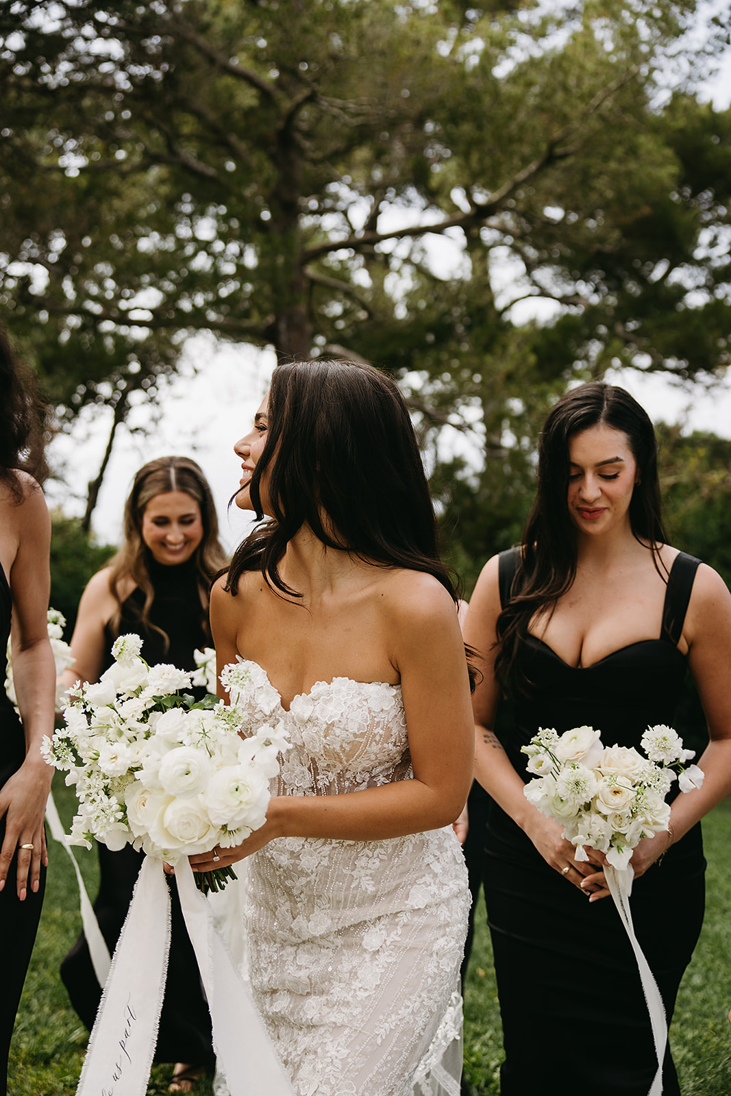 A bride stands with five bridesmaids, each holding white flower bouquets. The women are dressed in black dresses, and they are outdoors with trees and greenery in the background.