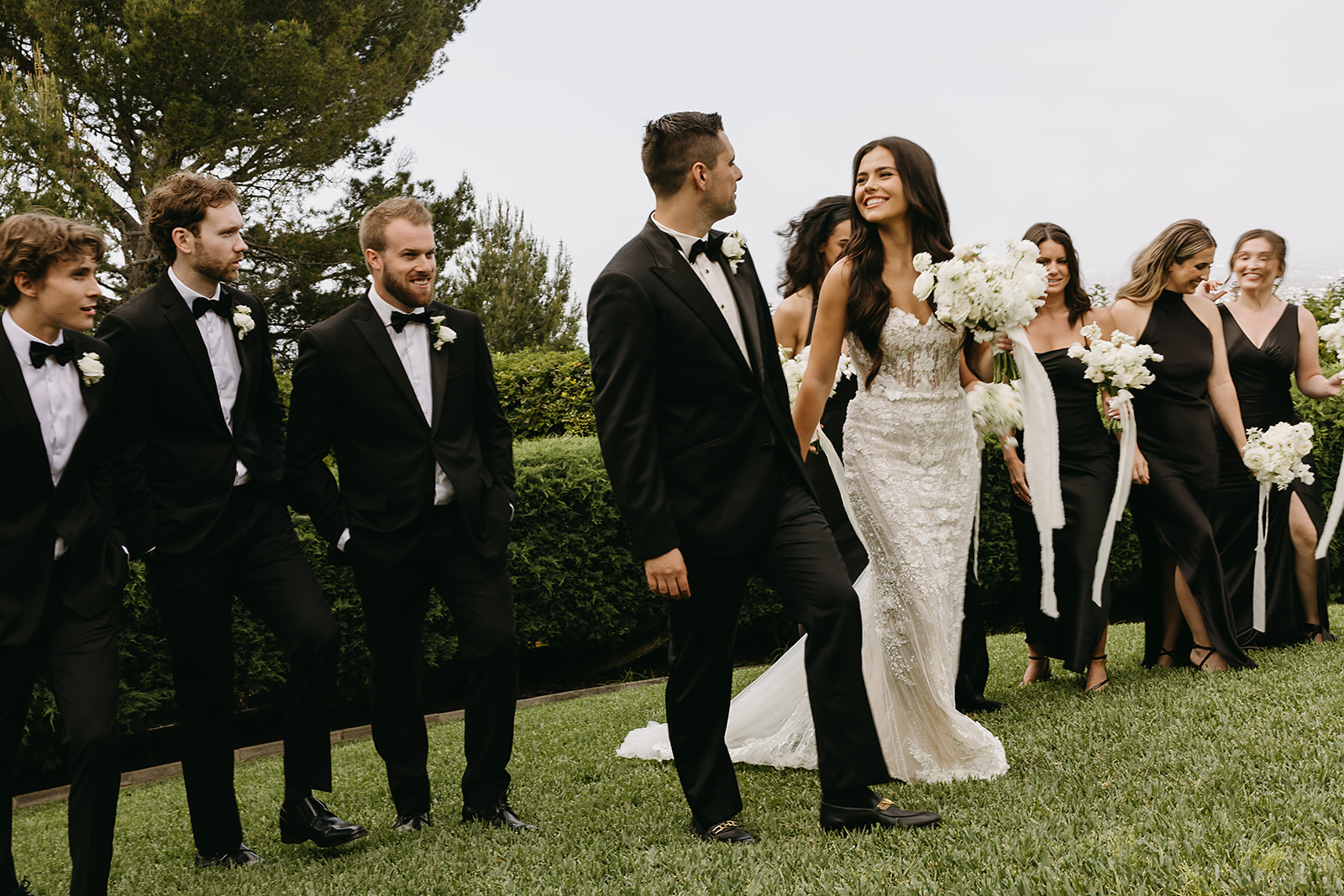 A bride and groom walk outdoors with their wedding party, who are dressed in black. They are holding white flowers, surrounded by lush greenery and trees.