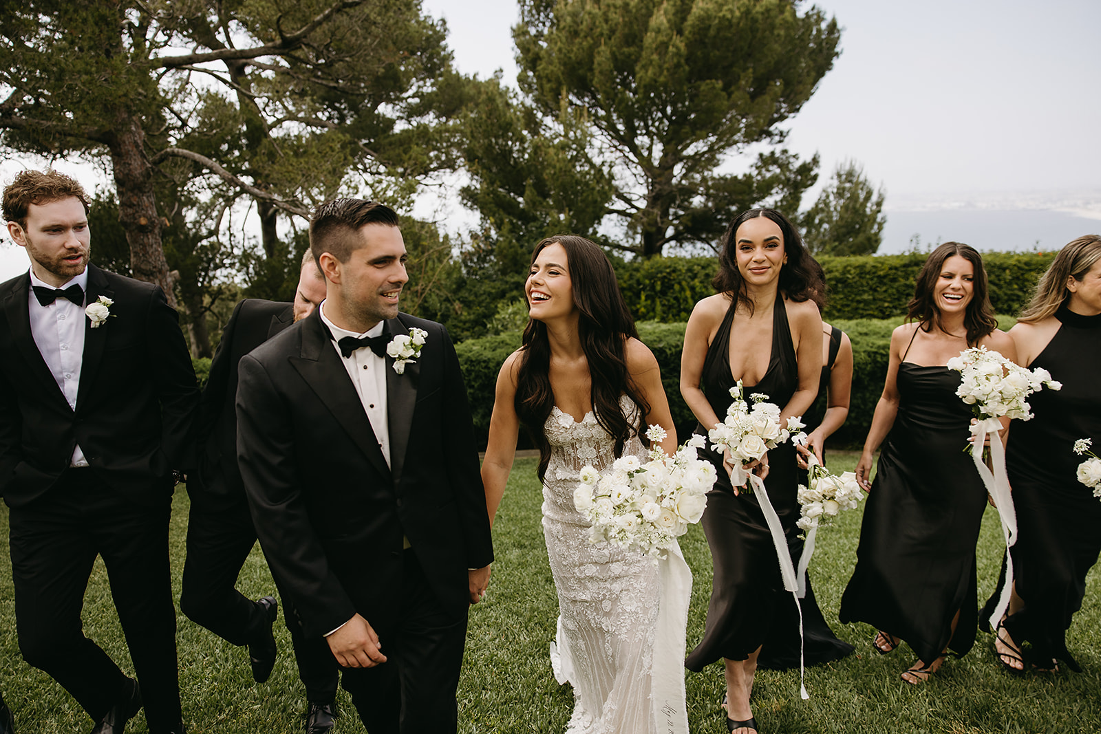 A bride and groom walk outdoors with their wedding party, who are dressed in black. They are holding white flowers, surrounded by lush greenery and trees.