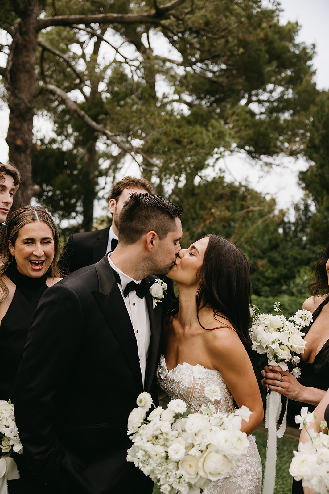 A bride and groom walk outdoors with their wedding party, who are dressed in black. They are holding white flowers, surrounded by lush greenery and trees.