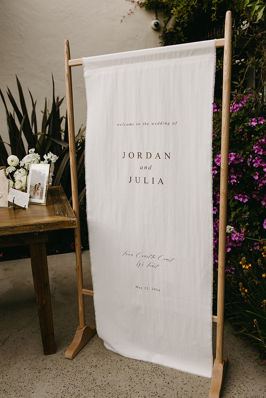 Wedding sign on a wooden stand set in a garden, with flowers in the background. The sign reads, "Welcome to the wedding of Jordan and Julia, Our favorite day of forever, March 3, 2022.