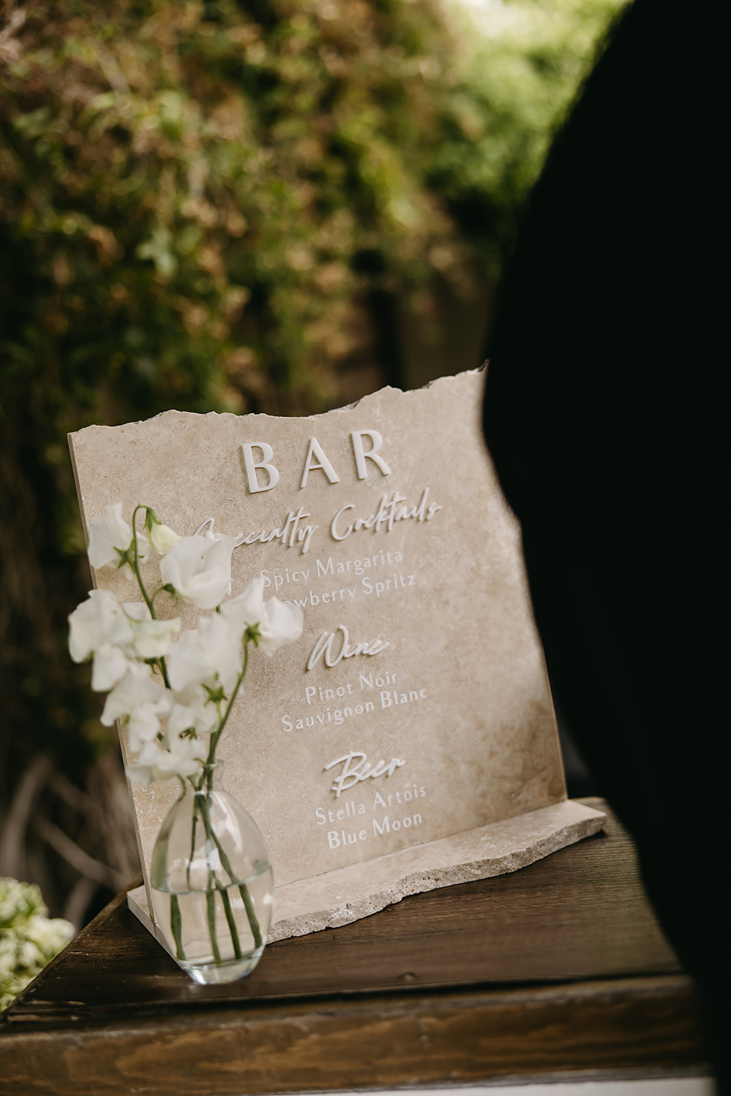 A bar menu displayed on a textured board with a small vase of white flowers beside it.