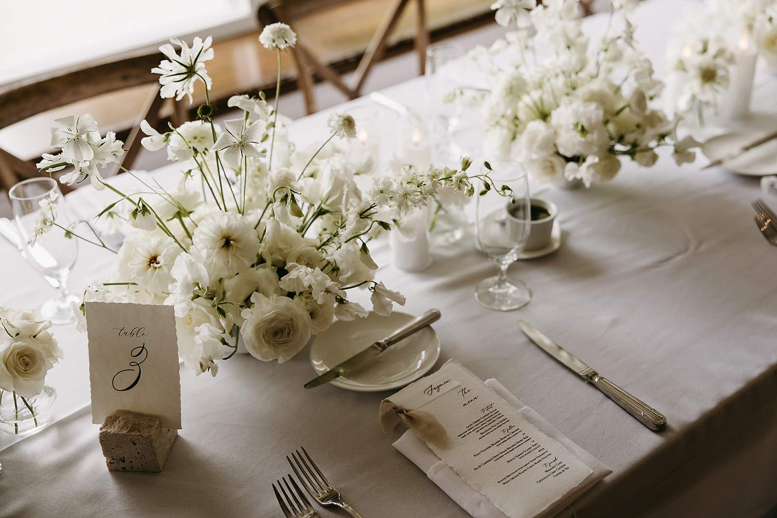 Elegant dining table set for an event with a white tablecloth, floral arrangements, and wooden chairs in a room with large windows and a view of greenery outside.