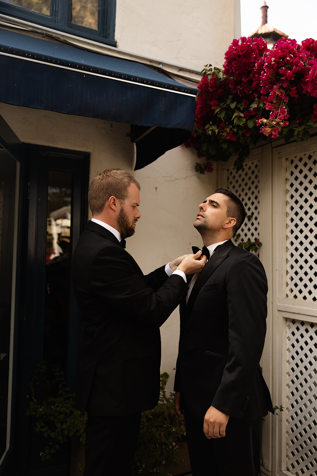 Two men in suits stand outside near a wall with flowers. One man adjusts the other's tie.