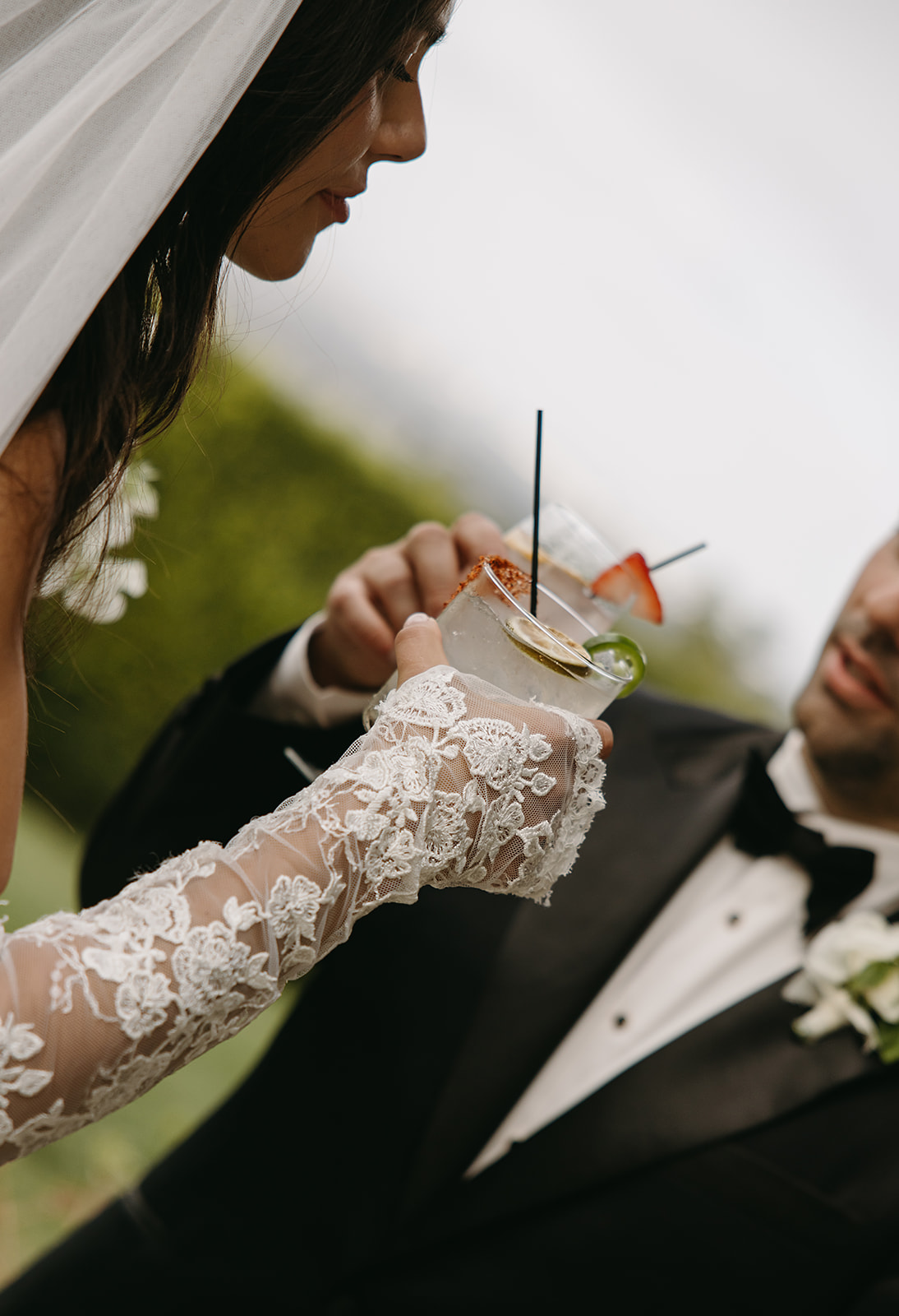 A bride and groom clink glasses outdoors. The bride wears lace gloves, and the groom is in a tuxedo with a boutonniere.