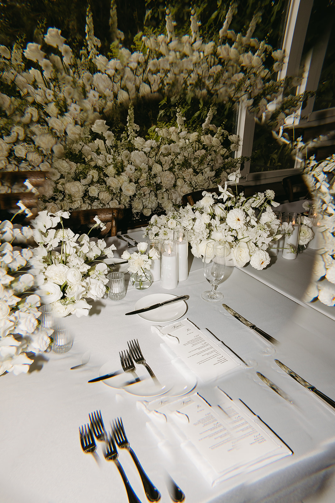 A white table setting with neatly arranged silverware and menus surrounded by abundant white flower arrangements.