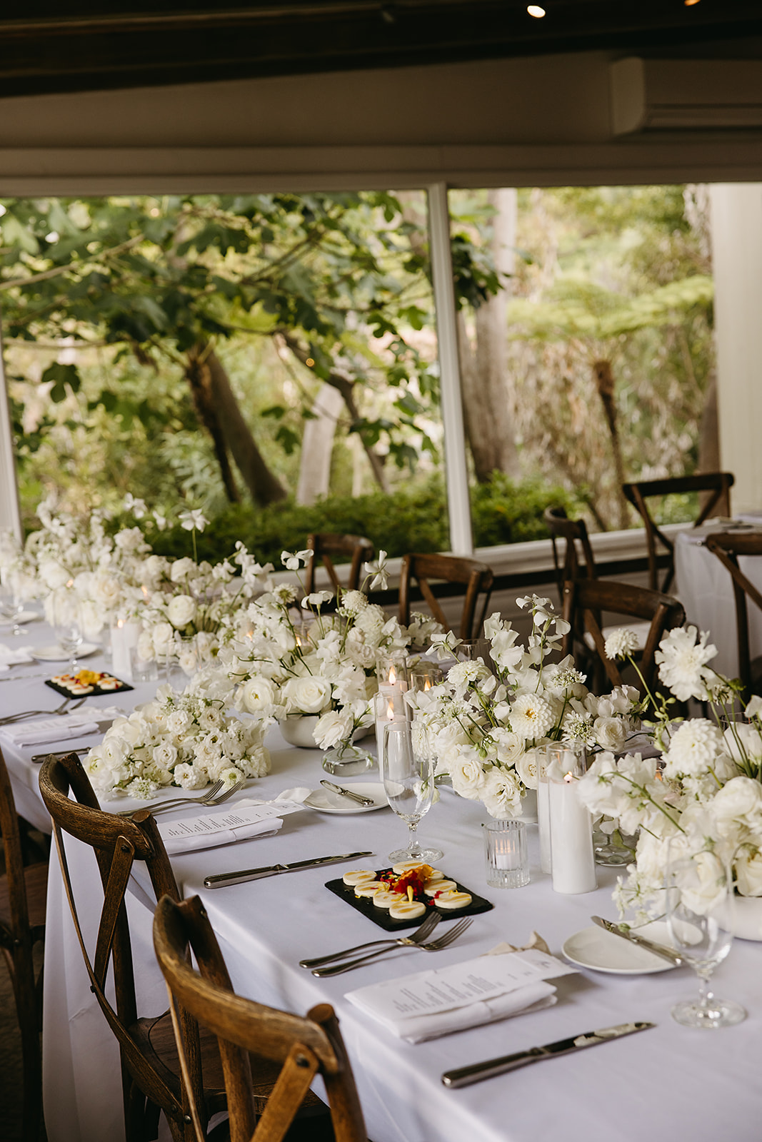 Elegant dining table set for an event with a white tablecloth, floral arrangements, and wooden chairs in a room with large windows and a view of greenery outside.