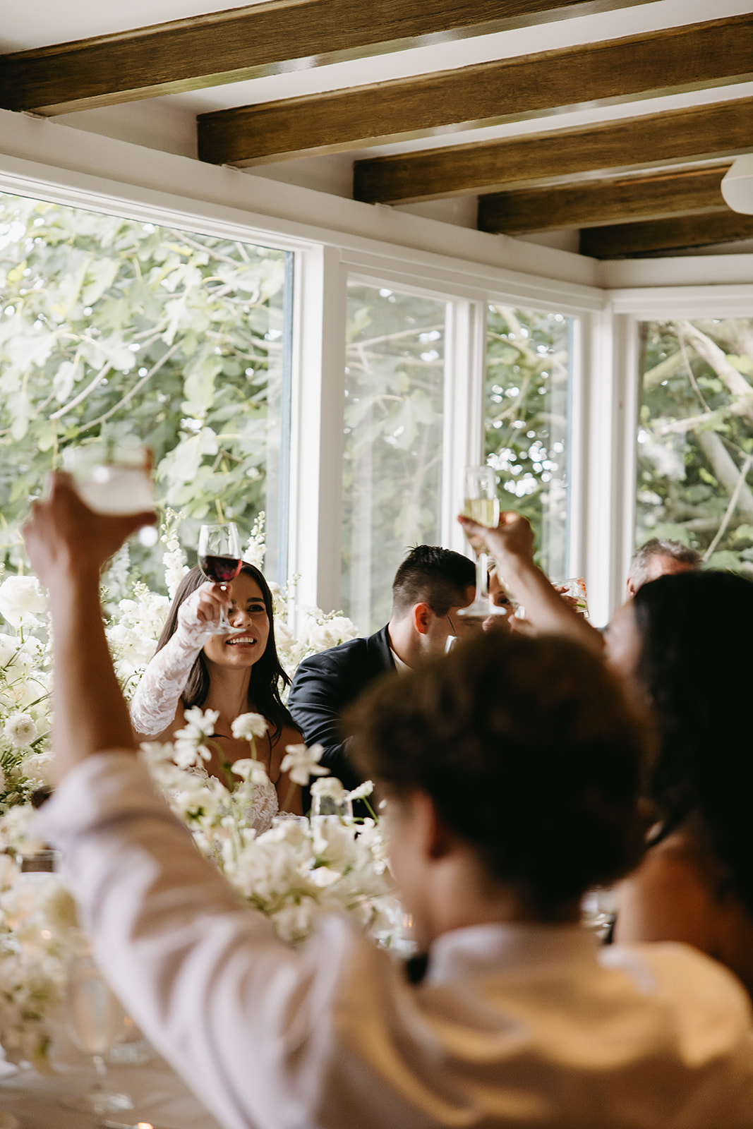 People seated at a dining table raise their glasses in a toast, surrounded by white floral arrangements, with large windows providing a view of greenery outside.