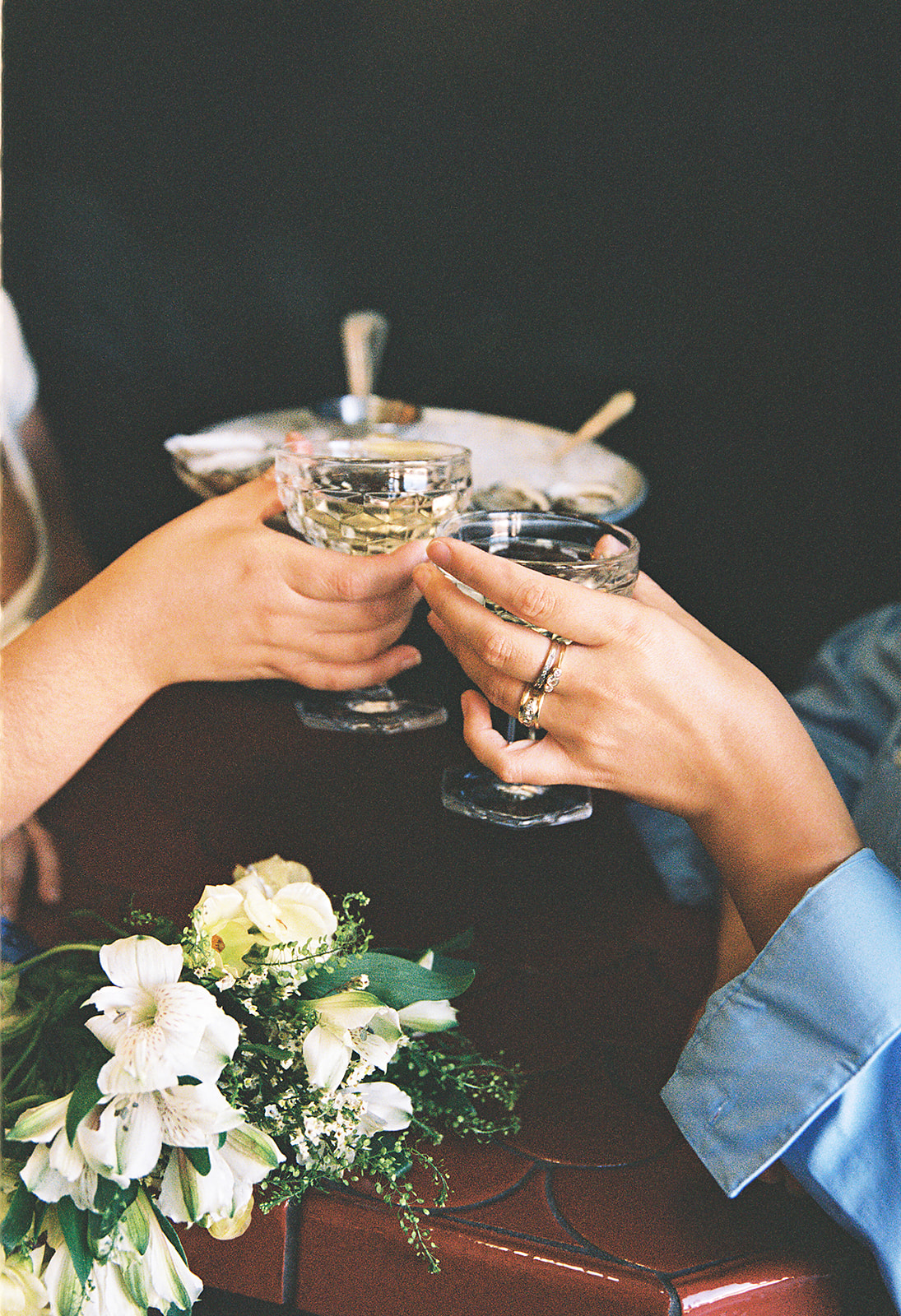 Two people clink glasses in a toast at a table, with a bouquet of white flowers and a dish in the background.