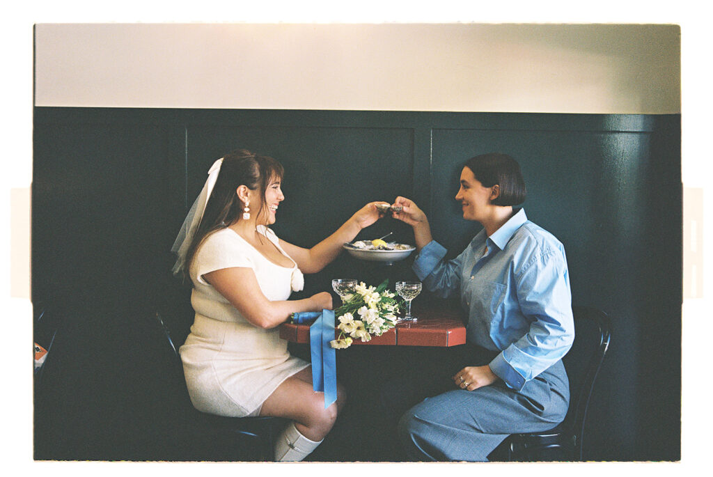 Two people clink glasses in a toast at a table, with a bouquet of white flowers and a dish in the background.