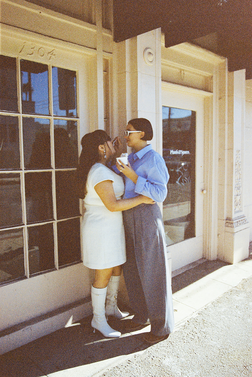 A person in a white dress and boots embraces another wearing a blue shirt and gray pants, standing outside a building with glass doors for their romantic engagement photos in LA