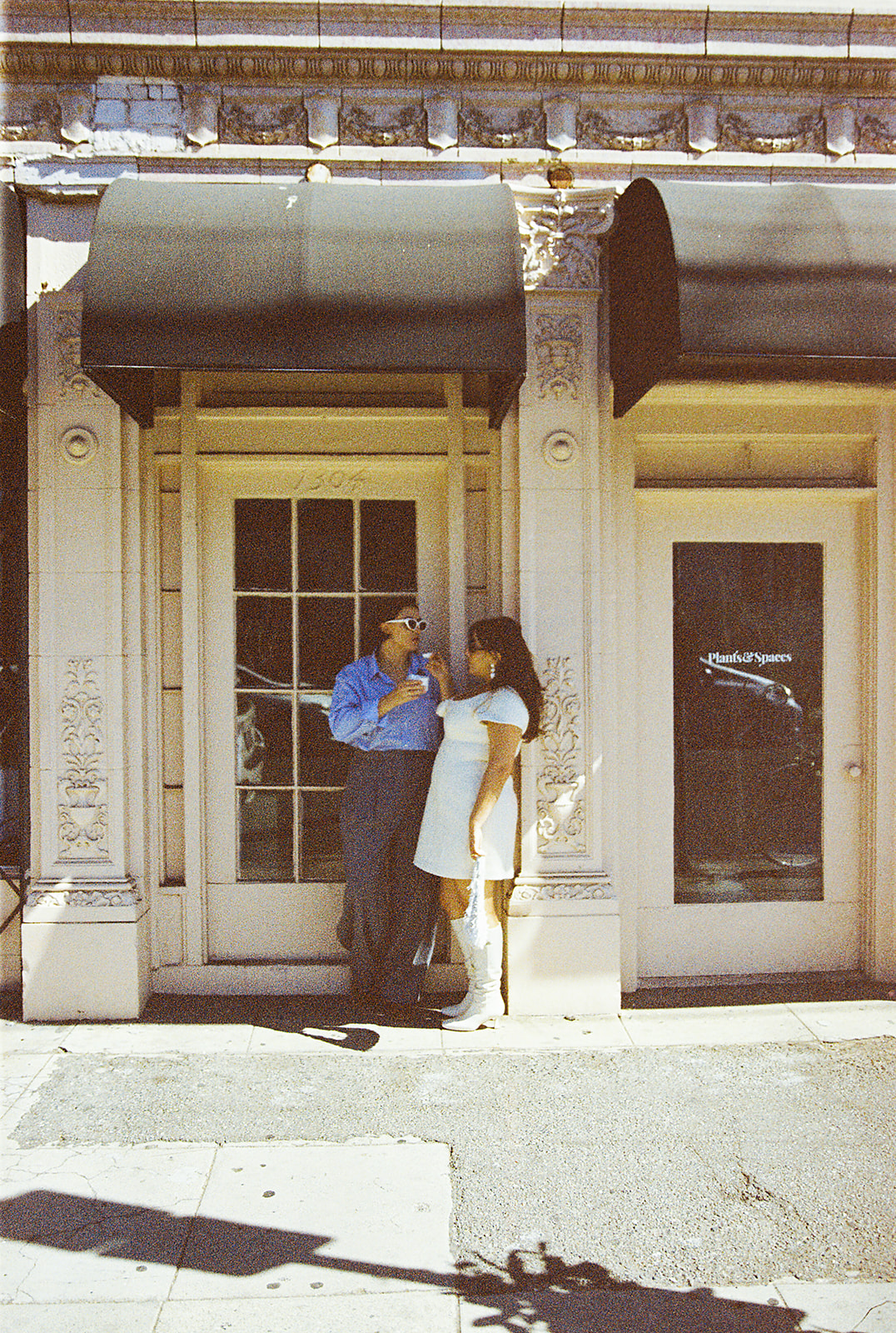 A person in a white dress and boots embraces another wearing a blue shirt and gray pants, standing outside a building with glass doors for their romantic engagement photos in LA