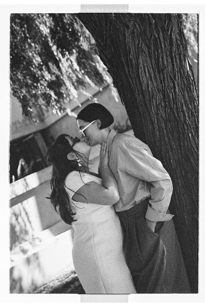 A couple shares a kiss outdoors on a sunny day, surrounded by greenery and trees.