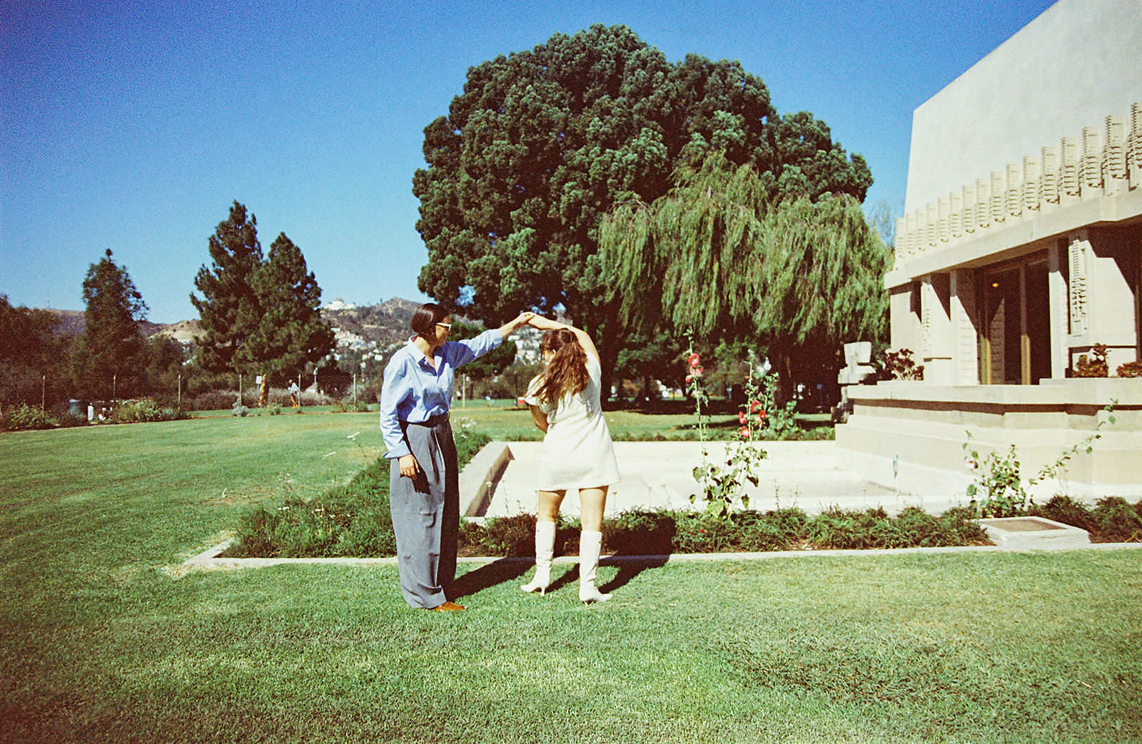 Two people dancing on a lawn in a sunny garden, surrounded by trees and a building on the right.