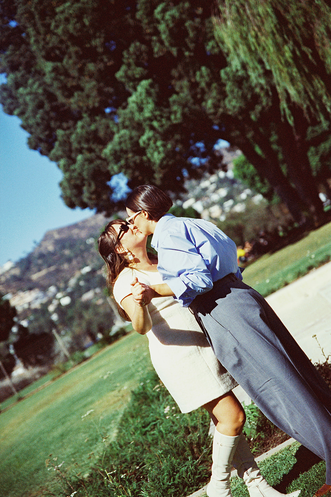 A couple kisses in a park with large trees and hills in the background. They are standing together, holding hands.